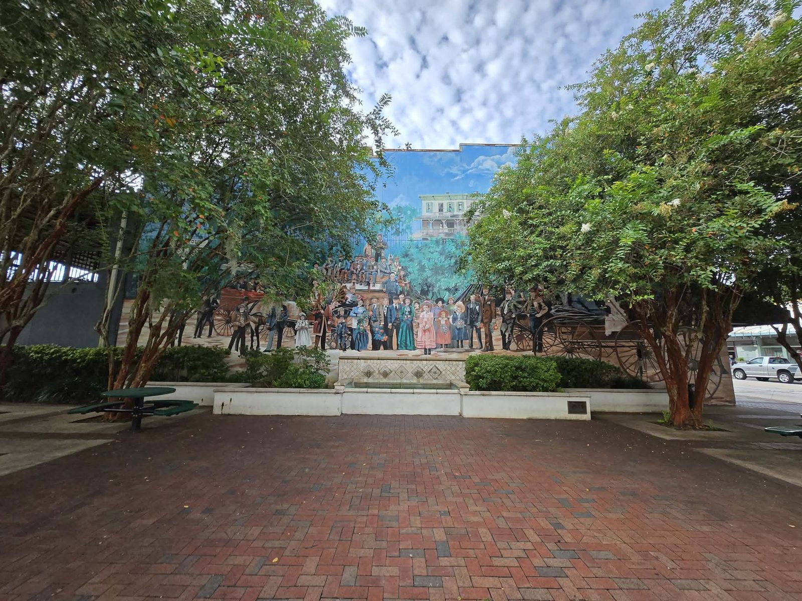 Mural of Victorian-era DeLand townspeople and horse-drawn carriages on a wall at a tree-shaded brick pocket park.