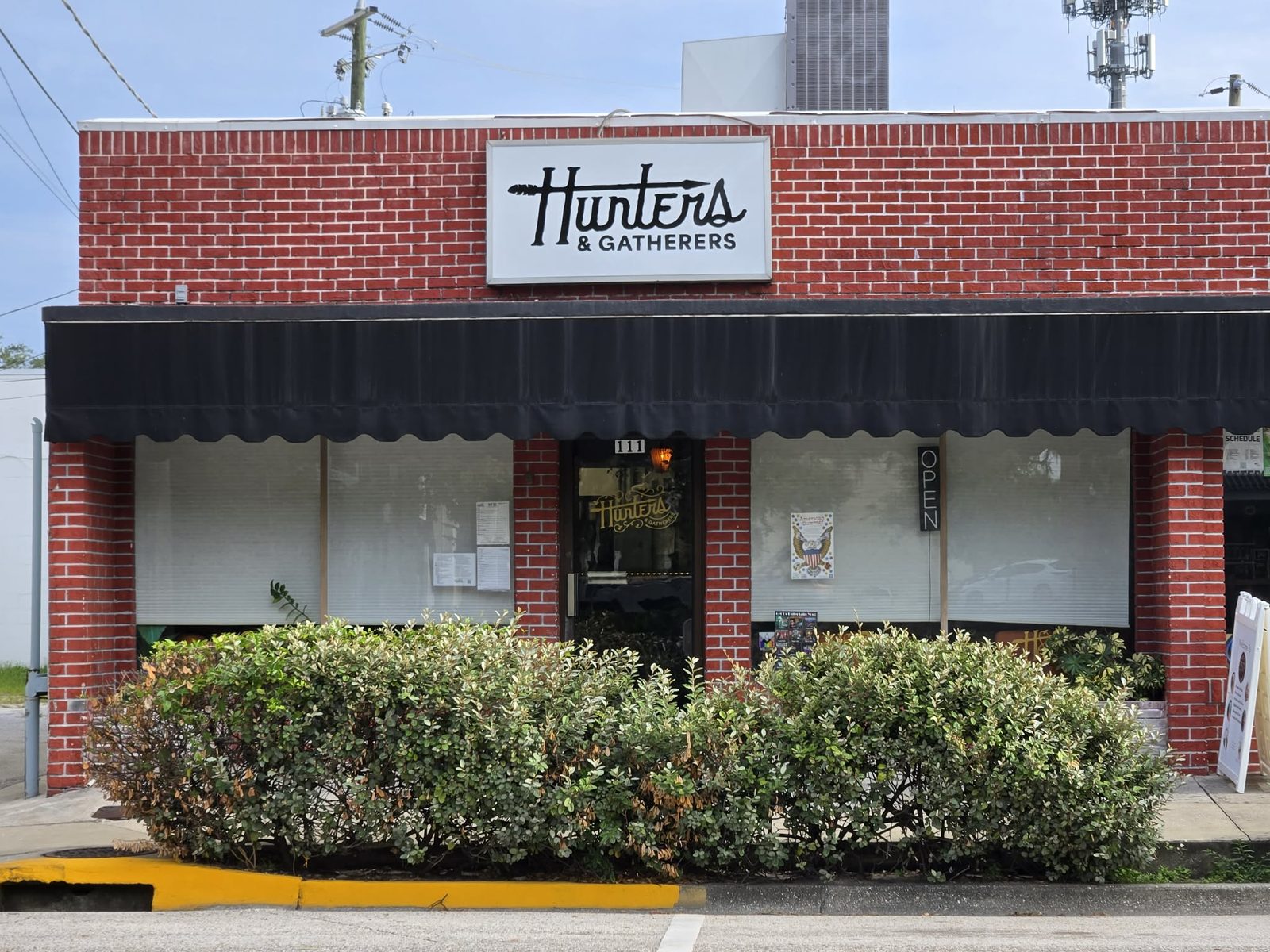 Red brick storefront of Hunters and Gatherers with a black awning and hedges in downtown DeLand.