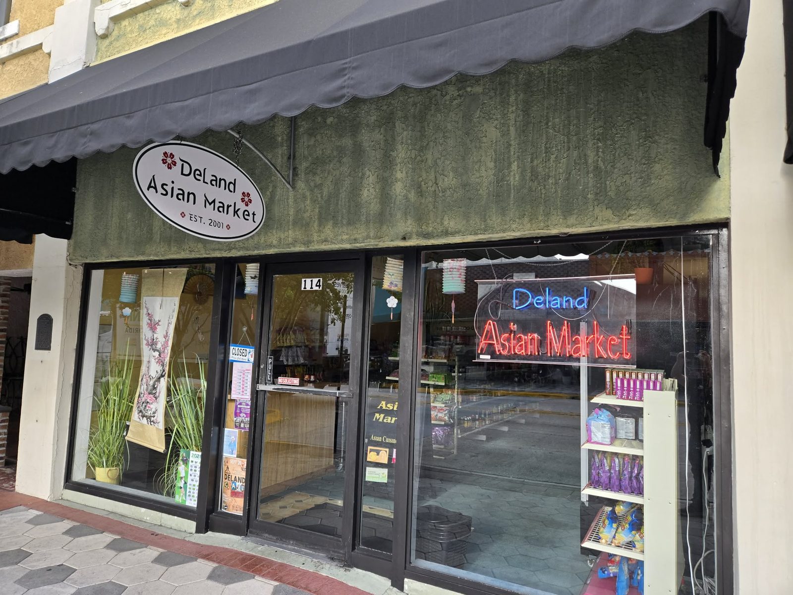 Storefront of DeLand Asian Market with an oval hanging sign and neon window sign in downtown DeLand.